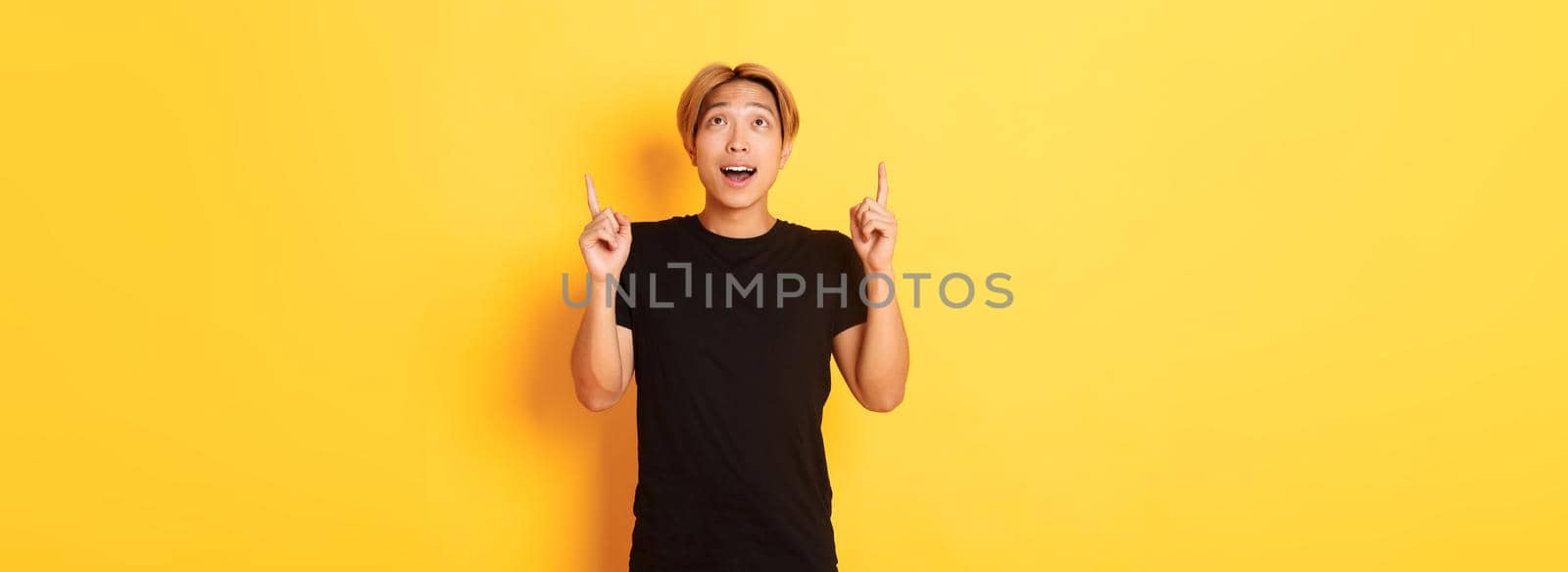 Impressed good-looking asian blond guy, wearing black t-shirt, looking and pointing fingers up excited, yellow background by Benzoix