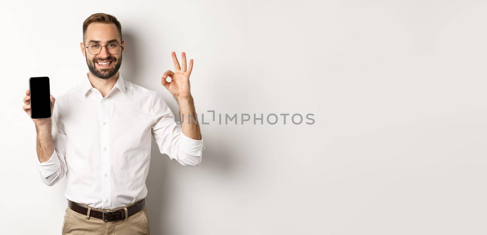 Satisfied young manager showing smartphone screen and okay sign, recommending application, standing over white background by Benzoix