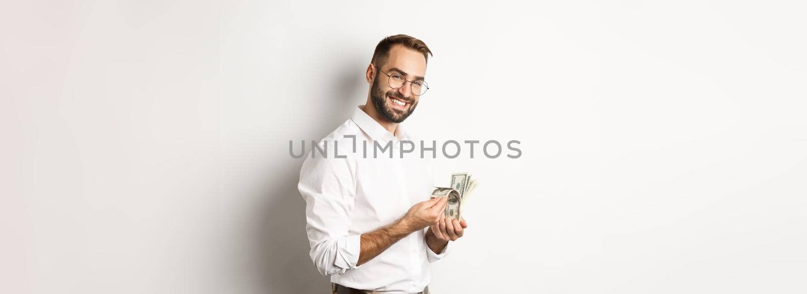 Successful business man counting money and smiling, standing against white background and looking satisfied.