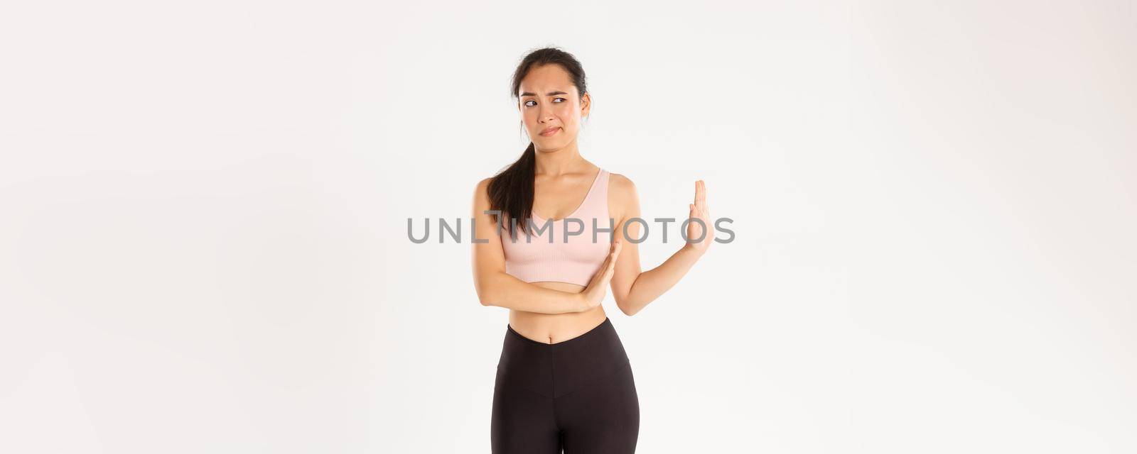 Sport, wellbeing and active lifestyle concept. Displeased and unamused asian fitness girl showing block with hand and looking left disgusted, express aversion and reject something bad.