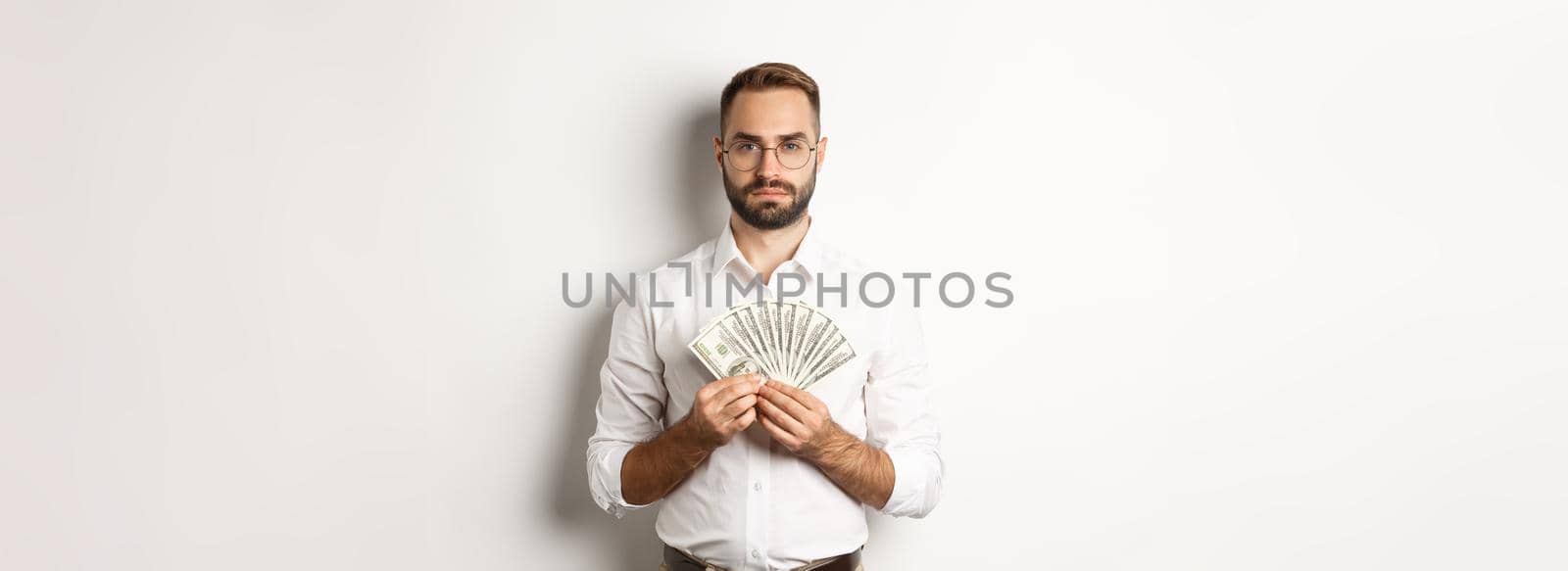 Serious businessman holding money, showing dollars, standing over white background by Benzoix