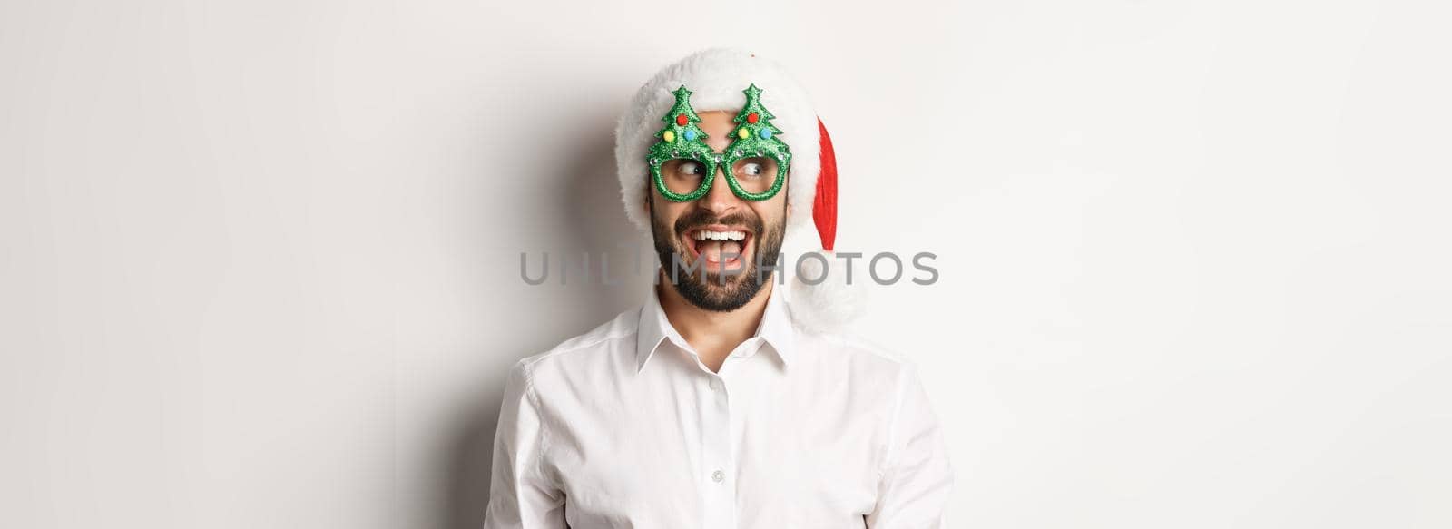 Close-up of funny man looking left with surprised face, wearing christmas party glasses and santa hat, celebrating New Year, white background.