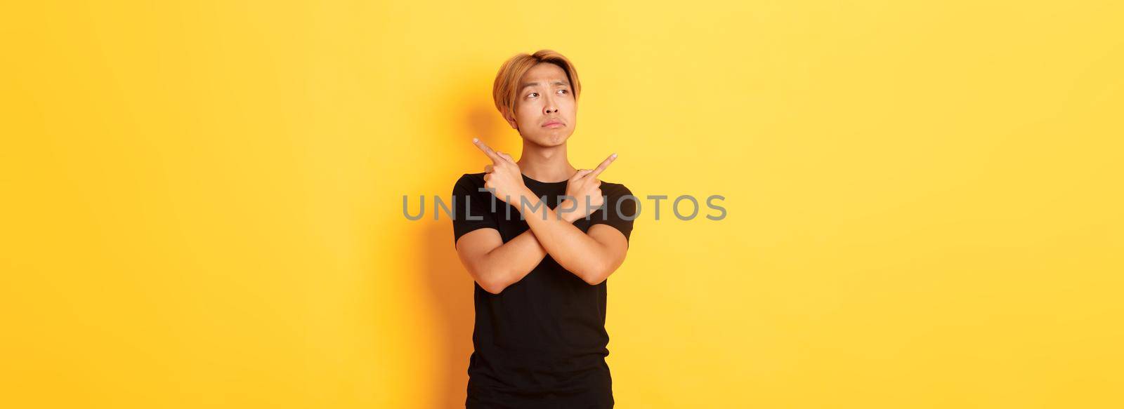 Indecisive handsome asian guy looking confused, pointing fingers sideways, standing yellow background by Benzoix