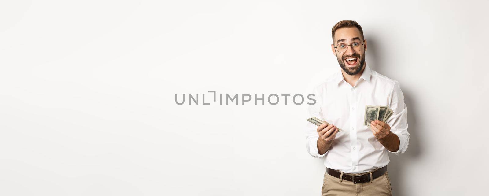 Handsome business man looking excited while counting money, standing over white background.