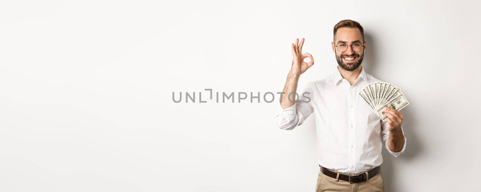 Satisfied young businessman showing money, make okay sign, earning cash, standing over white background.