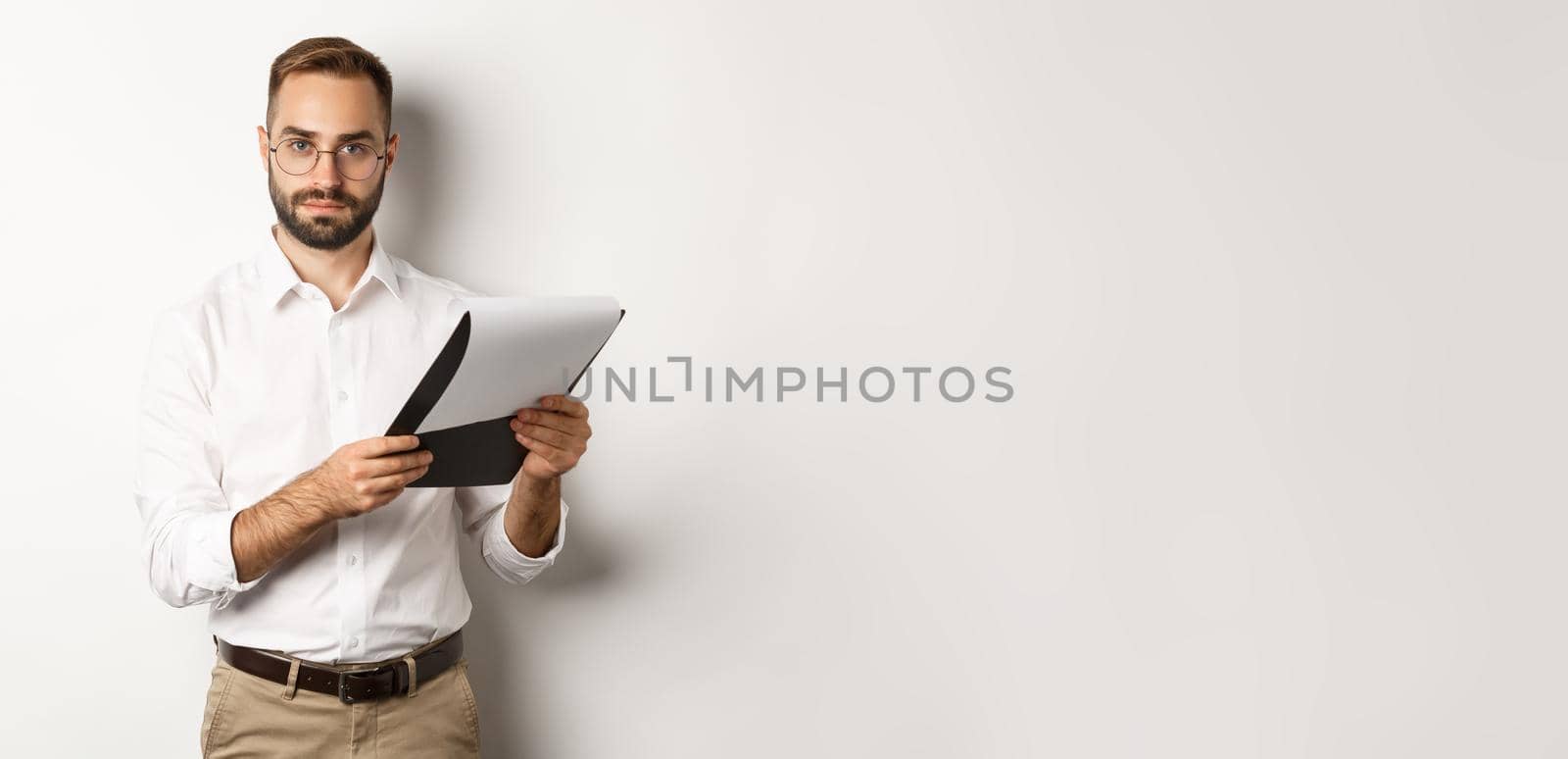 Serious employer looking at camera while reading documents on clipboard, having job interview, standing over white background by Benzoix