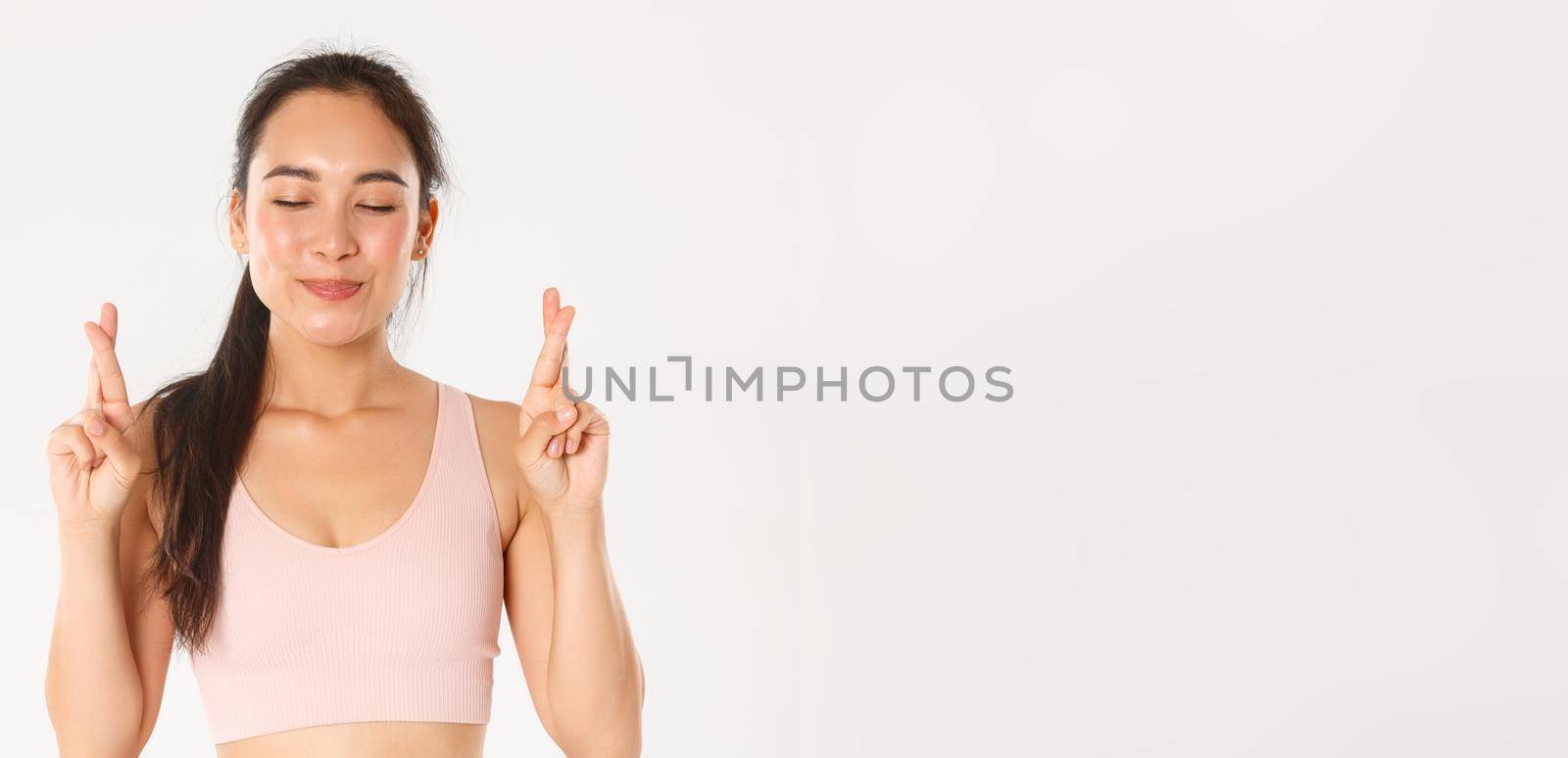 Sport, wellbeing and active lifestyle concept. Close-up of smiling optimistic asian girl, hope lose weight, cross fingers for good luck and close eyes while making wish, white background by Benzoix