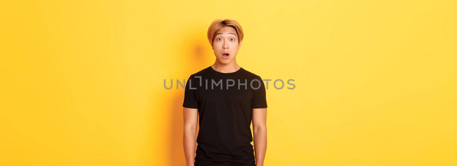 Portrait of startled handsome asian blond guy, drop jaw and gasping impressed, yellow background.