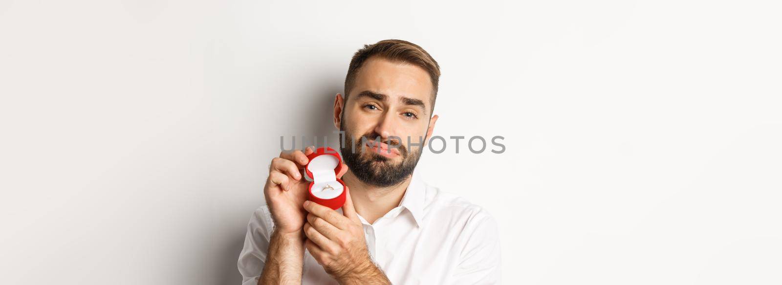 Close-up of hopeful man begging to marry him, looking sad and showing wedding ring, making a proposal, white background by Benzoix