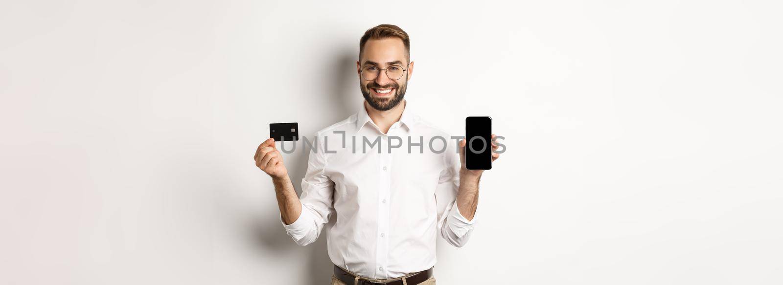 Business and online payment. Smiling handsome man showing mobile screen and credit card, standing over white background by Benzoix