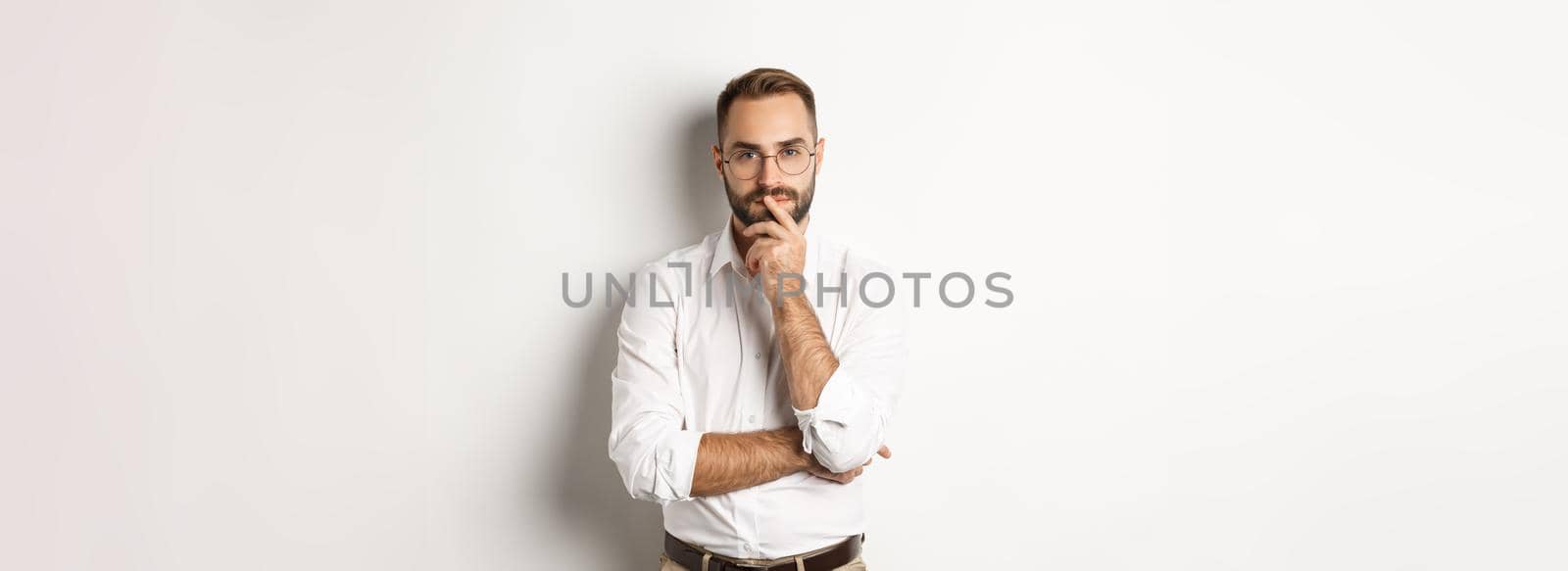 Thoughtful handsome businessman looking at camera, making choice or thinking, standing in glasses and white collar shirt against studio background.