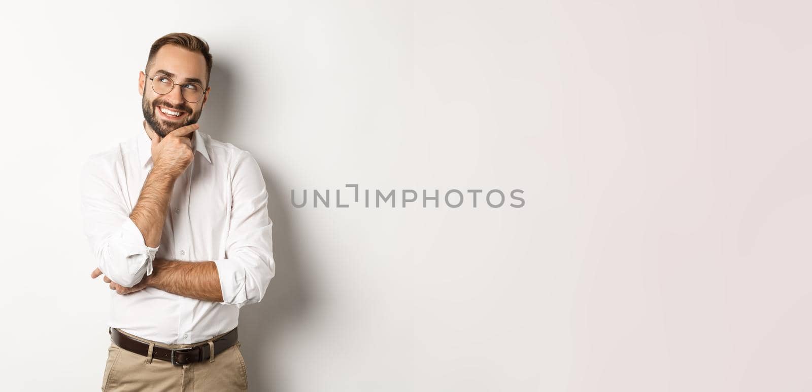 Satisfied businessman having interesting idea, looking at upper left corner and thinking with pleased smile, standing over white background.