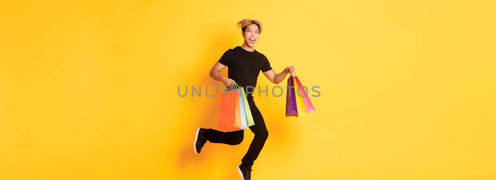 Full-length of happy attractive asian man jumping from happiness and carry shopping bags, standing yellow background.