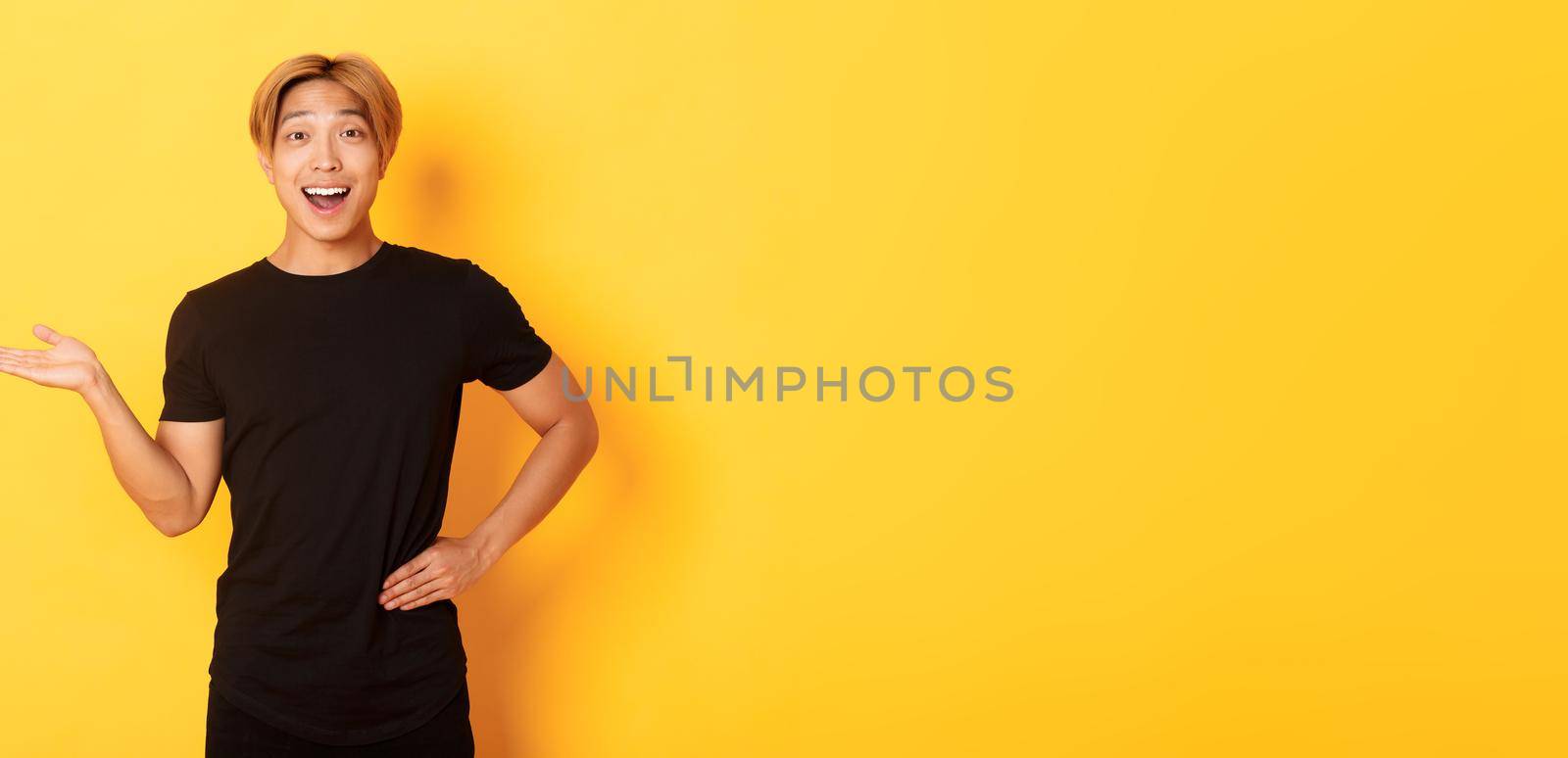 Portrait of happy and excited smiling asian guy holding something on hand over yellow background.