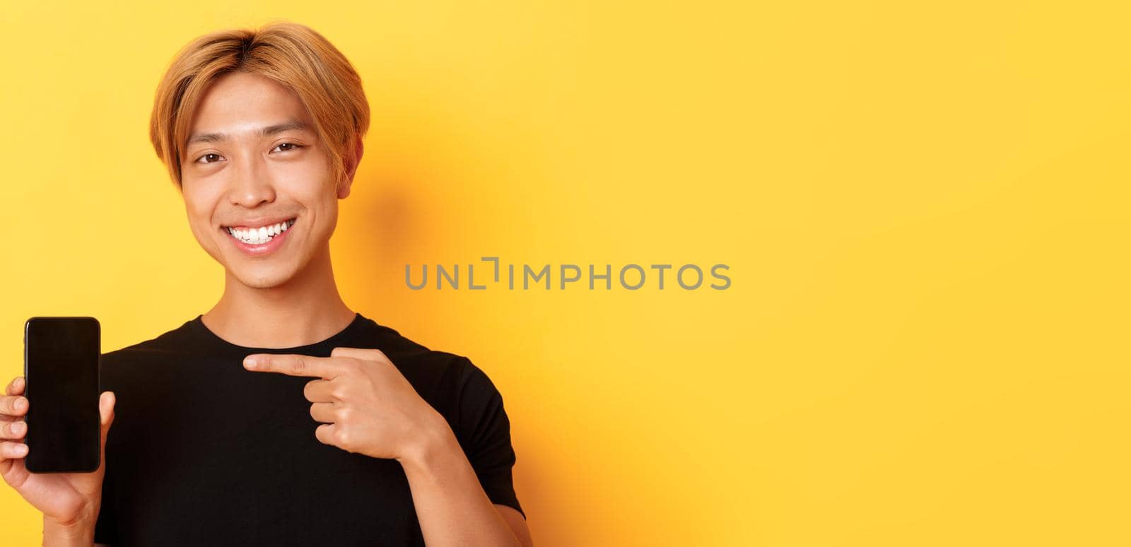 Cheerful attractive korean guy pointing finger at mobile phone screen, smiling pleased, standing over yellow background.