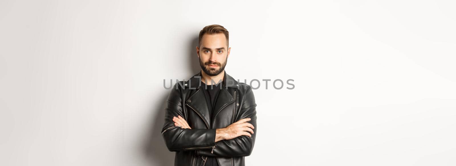 Attractive macho man in leather jacket smiling, looking confident with hands crossed on chest, white background.