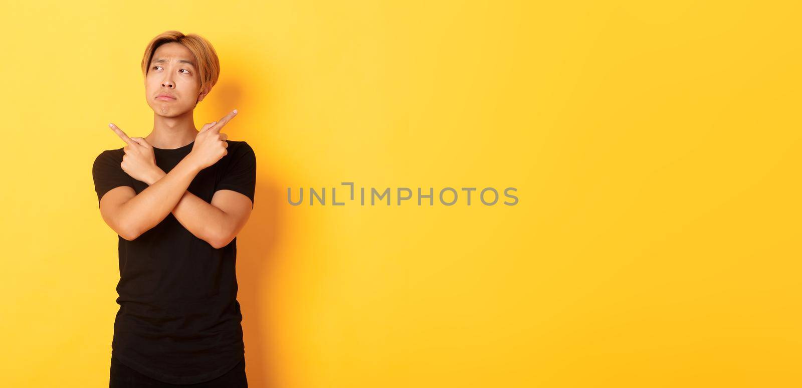 Indecisive handsome asian guy looking confused, pointing fingers sideways, standing yellow background.