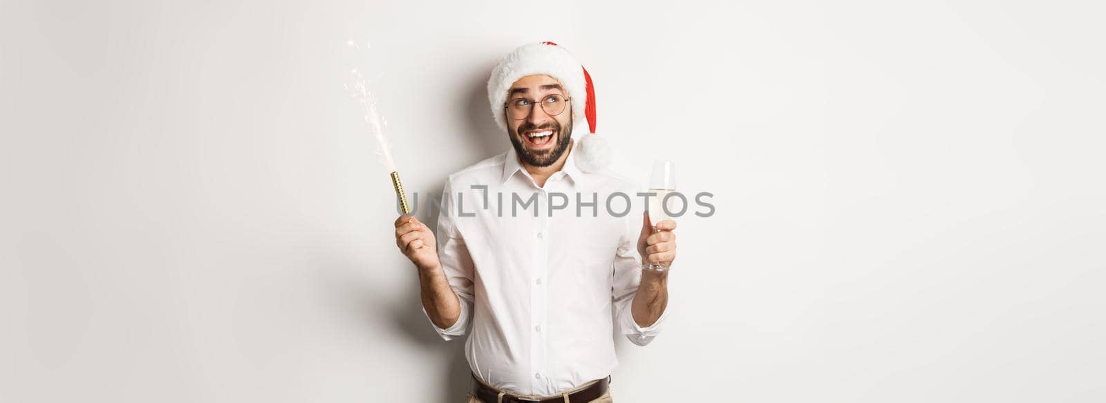 Winter holidays and celebration. Happy businessman enjoying New Year party, wearing Santa hat and drinking champagne, smiling amused, white background.