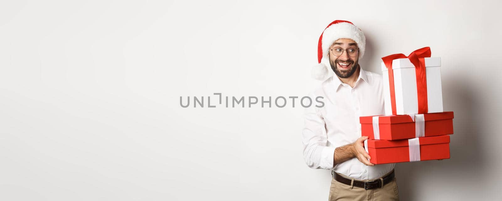 Merry christmas, holidays concept. Excited man celebrating xmas, wearing santa hat and holding gifts, standing over white background.