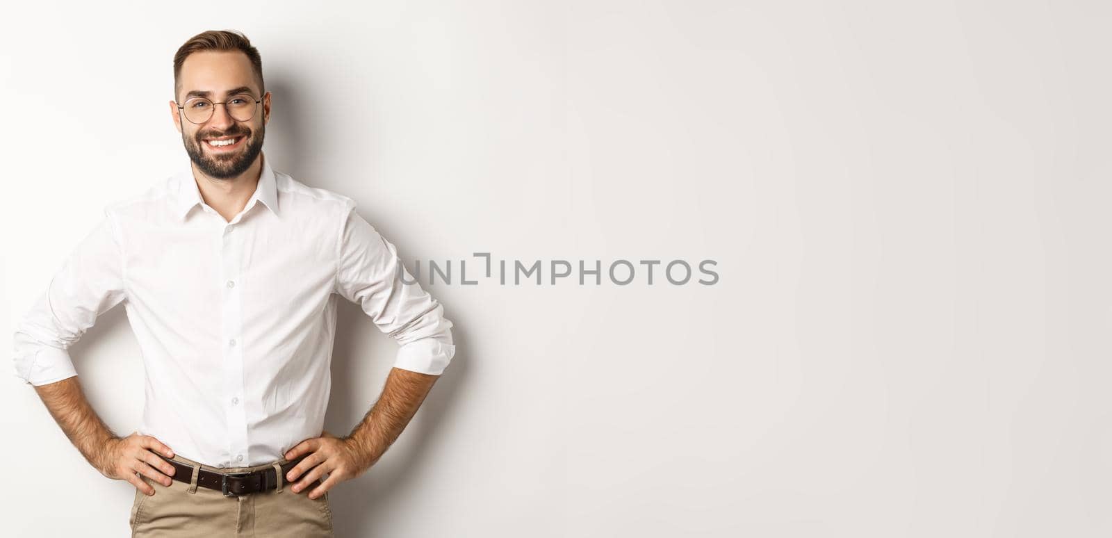 Handsome businessman in glasses looking satisfied, smiling and holding hands on waist, standing over white background.