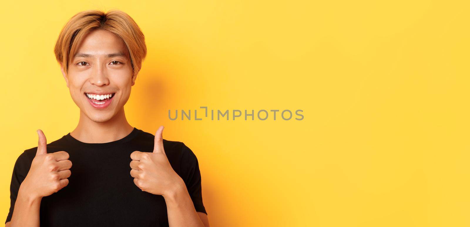 Close-up of satisfied, happy asian guy in black t-shirt, showing thumbs-up in approval, smiling and agree, standing yellow background.