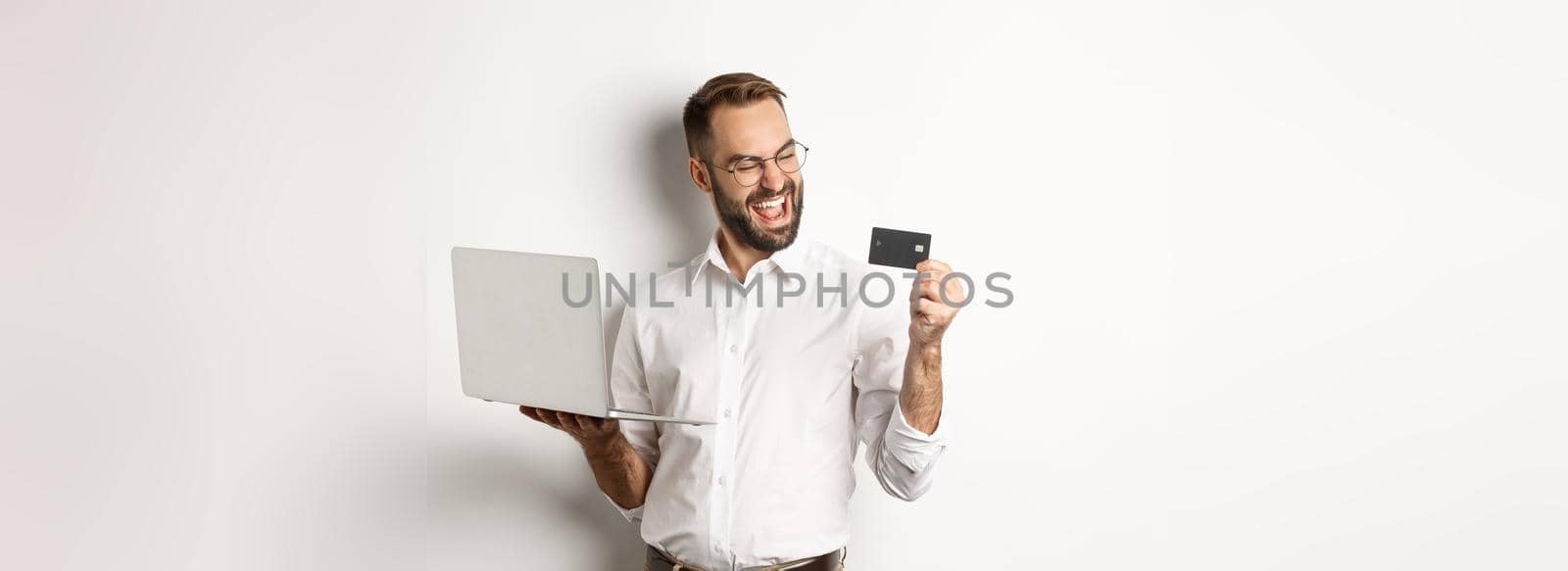Online shopping. Satisfied handsome man looking at credit card after making order internet, using laptop, standing over white background.