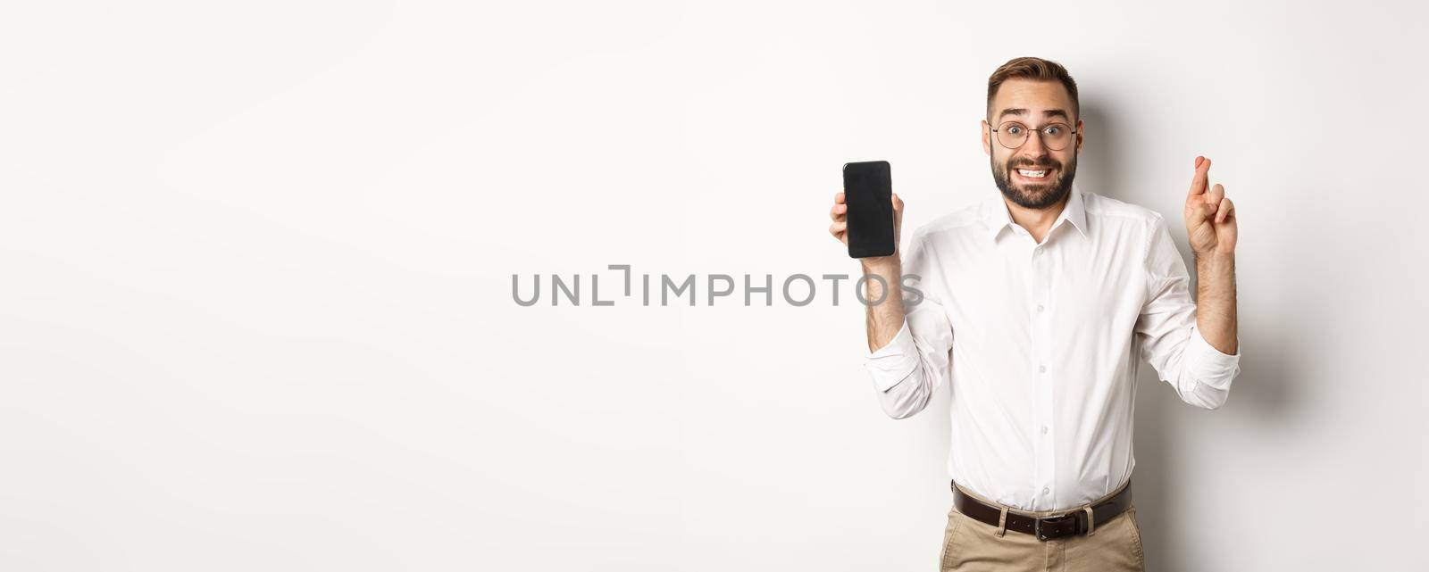 Hopeful young business man showing mobile screen, holding fingers crossed, waiting for online results, standing over white background.