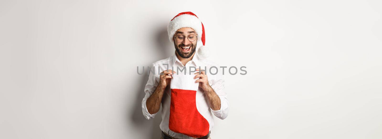 Happy adult man open christmas sock and looking inside, receiving xmas gifts for winter holidays, standing in santa hat, white background.