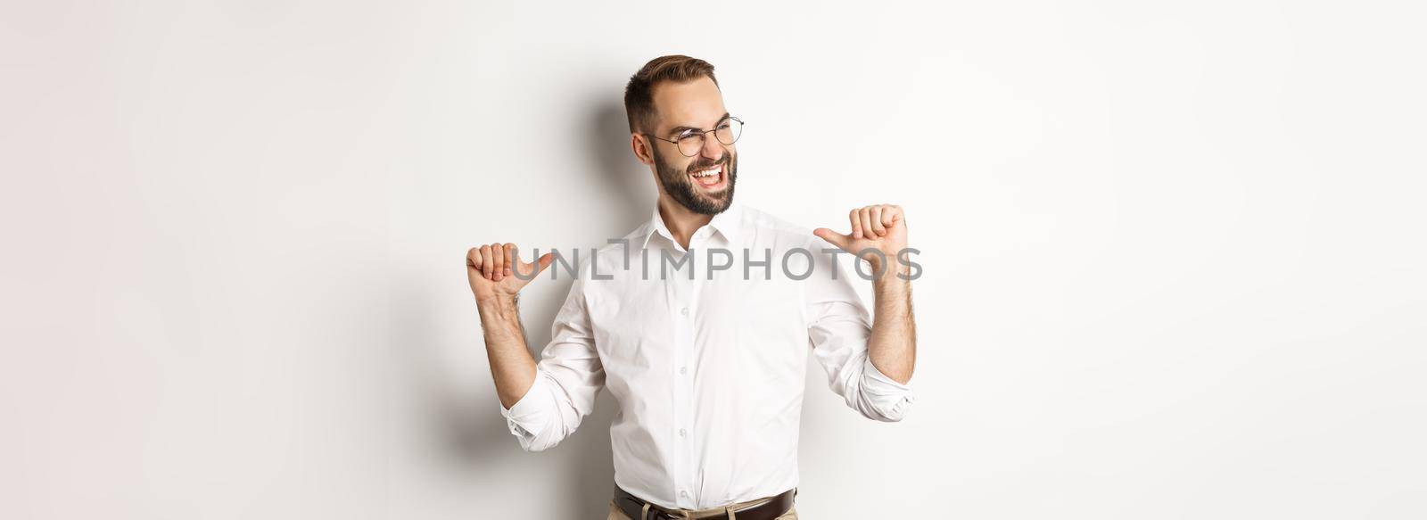 Satisfied and self-assured businessman pointing at himself, standing over white background.