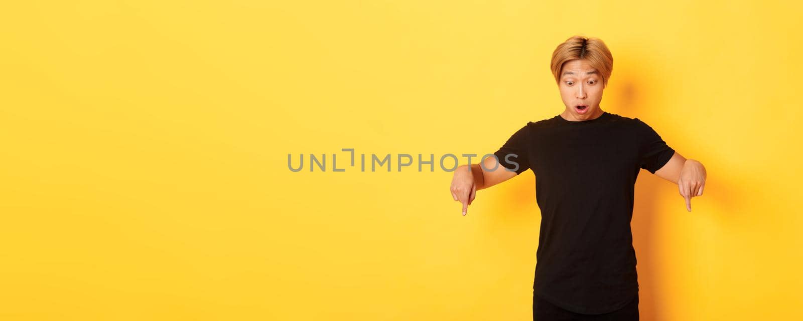 Portrait of surpirsed asian guy gasping amazed, pointing fingers and looking down at banner, standing yellow background by Benzoix