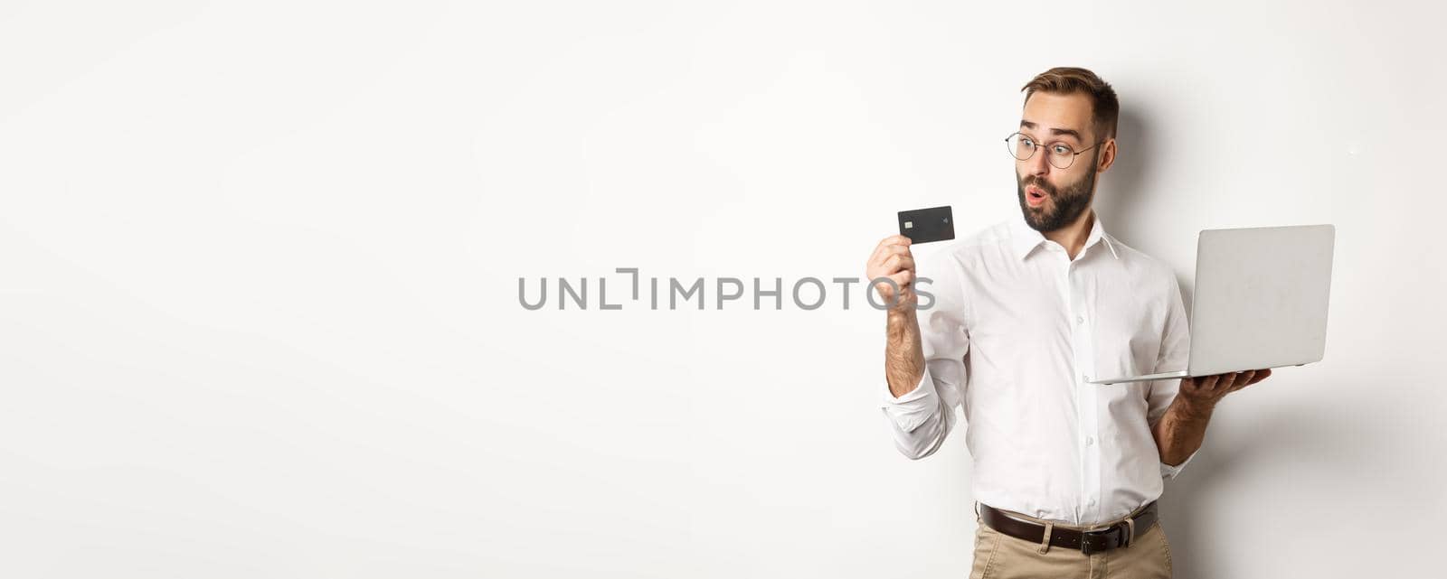 Online shopping. Amazed businessman holding laptop, looking impressed at credit card, standing over white background.