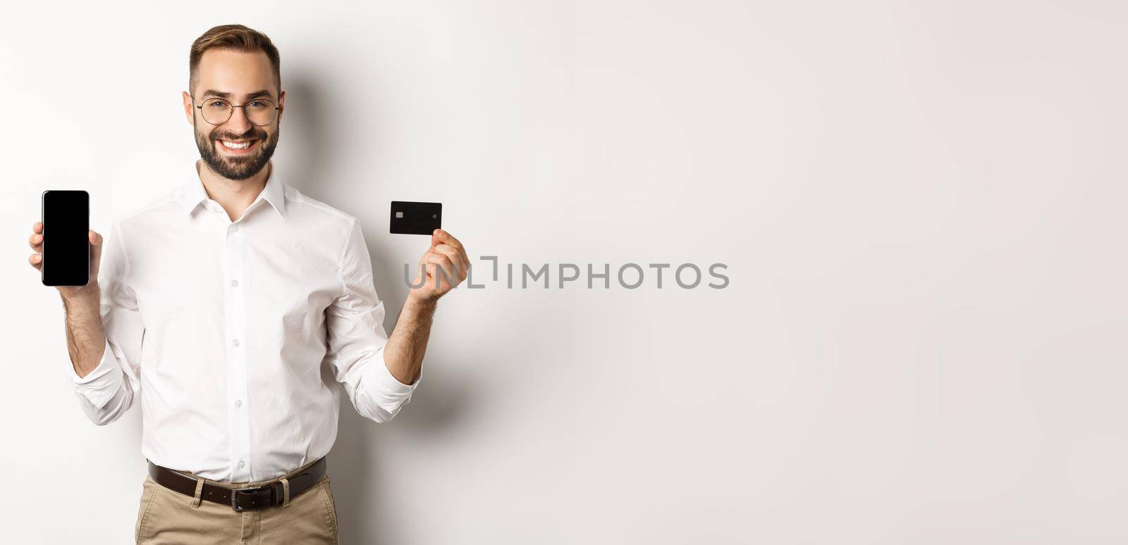 Business and online payment. Smiling handsome man showing mobile screen and credit card, standing over white background.