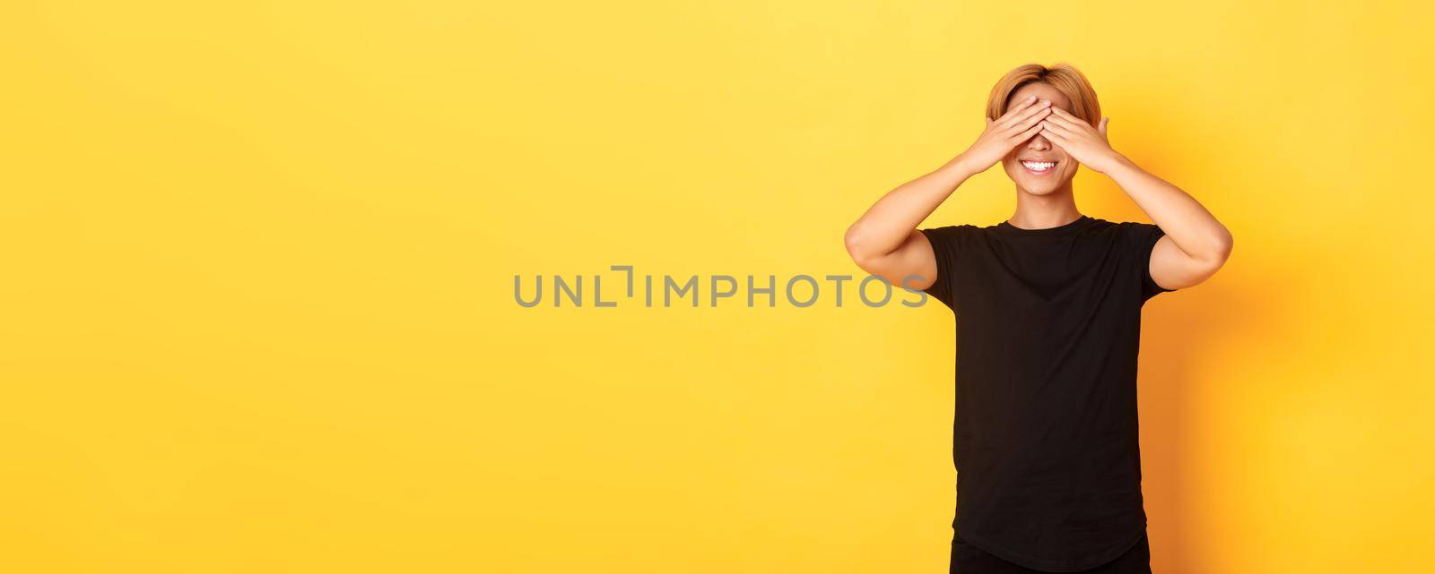 Portrait of excited happy asian guy, smiling and close eyes with hands, standing yellow background by Benzoix