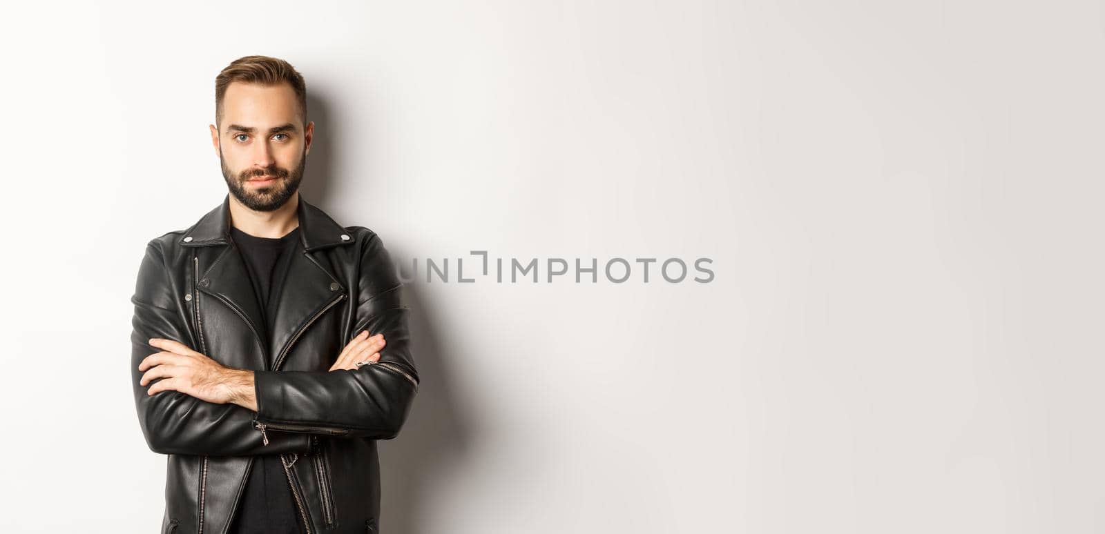 Attractive macho man in leather jacket smiling, looking confident with hands crossed on chest, white background.