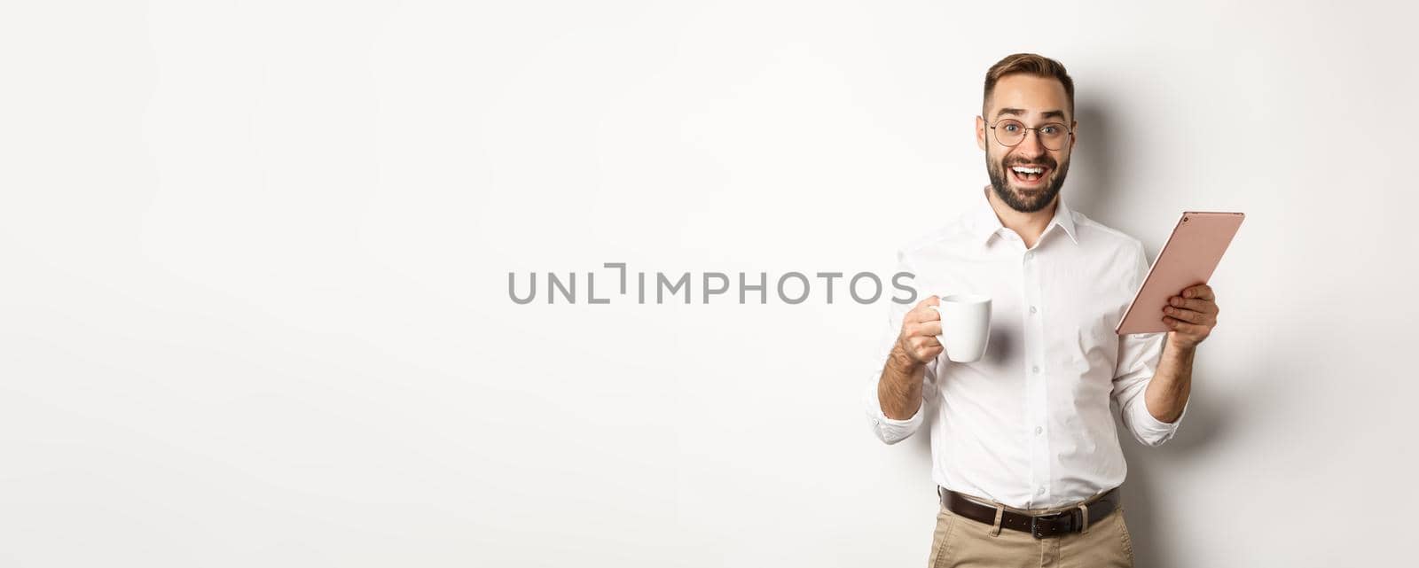 Excited manager reading on digital tablet, working and drinking coffee, standing against white background.