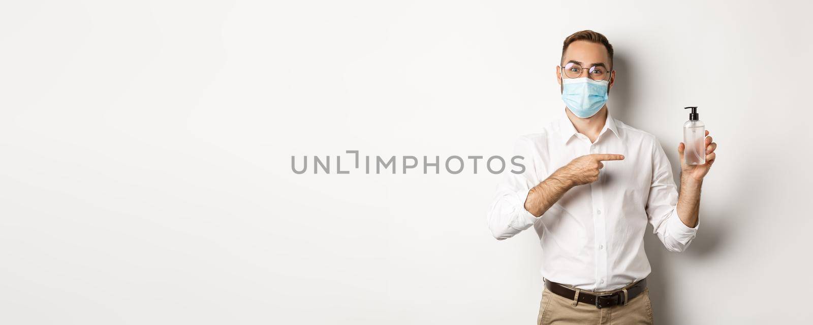 Covid-19, social distancing and quarantine concept. Office worker in medical mask pointing at hand sanitizer, showing antiseptic, white background.