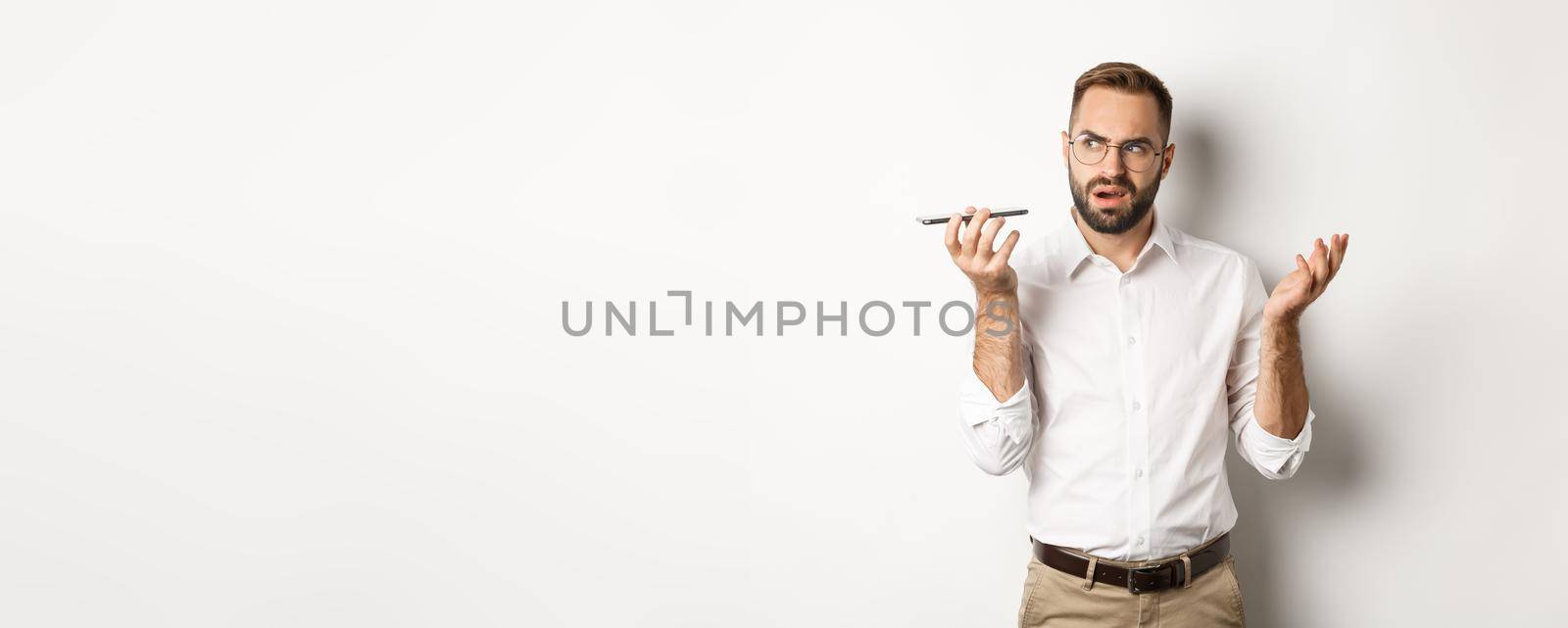 Man recording voice message or talking on speakerphone, looking confused, standing over white background.