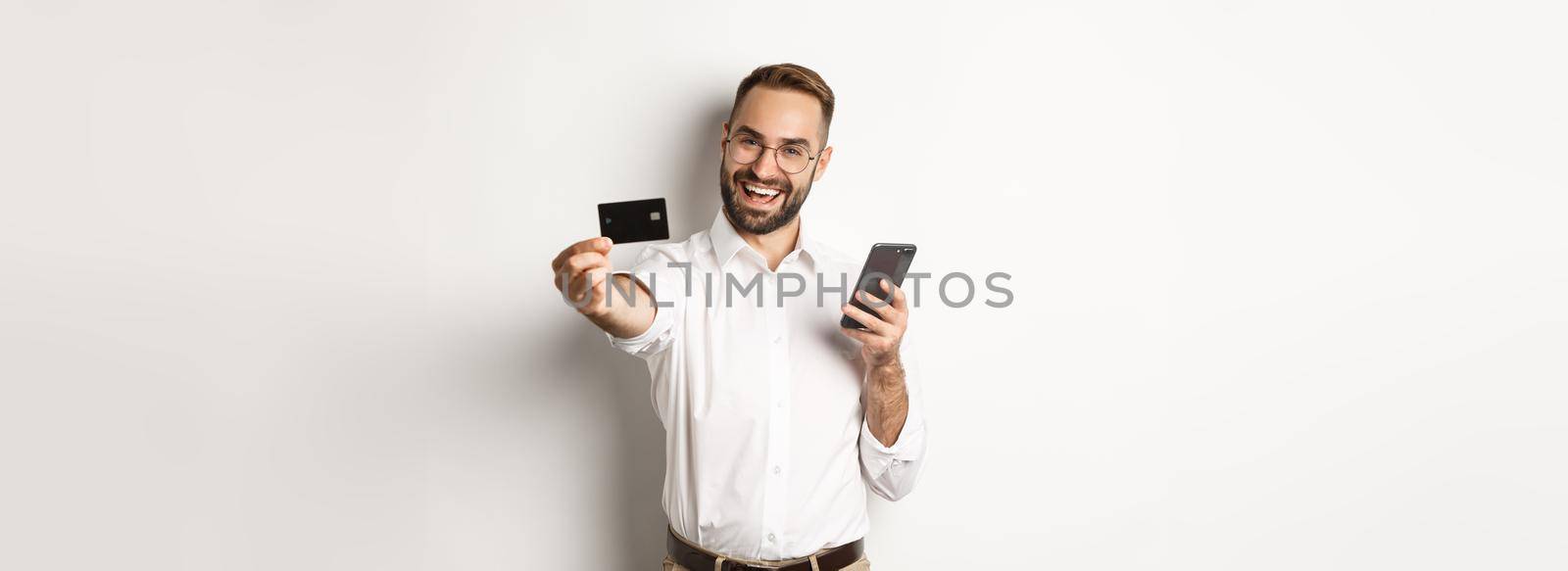 Business and online payment. Excited man showing his credit card while holding smartphone, standing satisfied against white background.