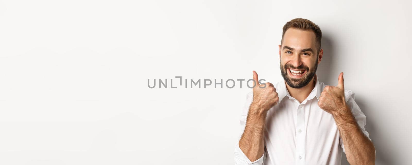 Close-up of satisfied bearded guy in white shirt, showing thumbs up in approval, like and agree, positive answer, white background by Benzoix