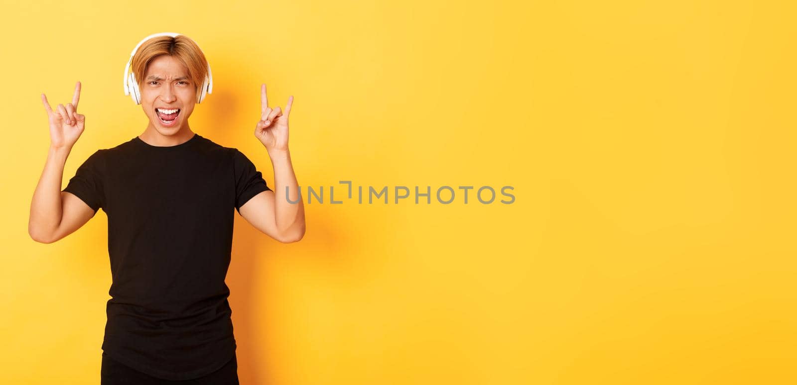 Joyful handsome asian guy enjoying listening music in headphones, showing rock-n-roll gesture, standing over yellow background by Benzoix