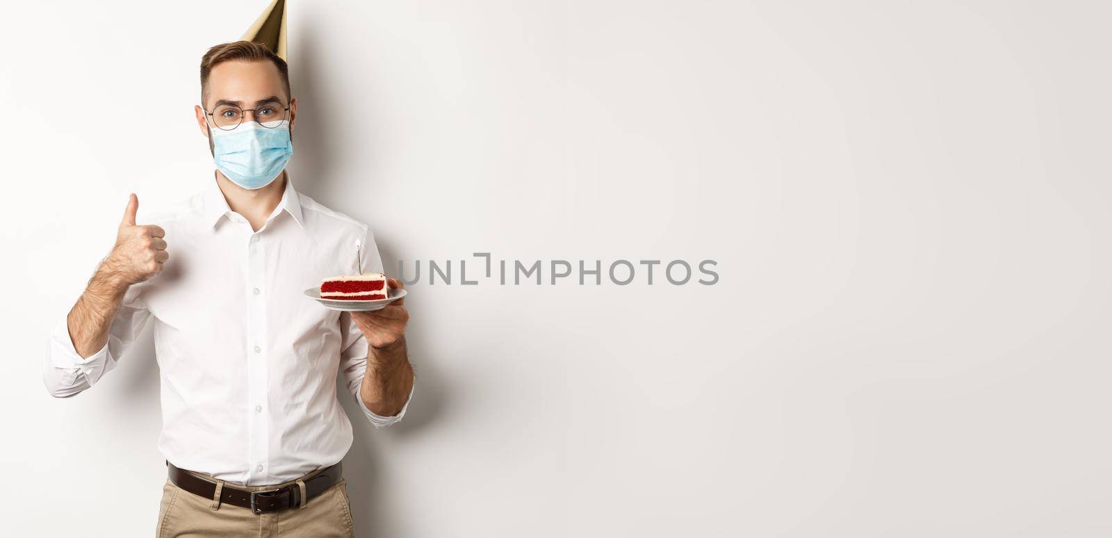Covid-19, social distancing and celebration. Happy young man in face mask, enjoying birthday party, holding bday cake and make thumb up gesture, white background.