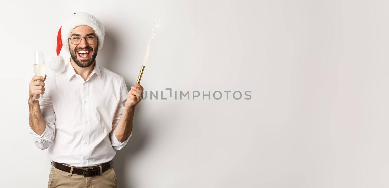 Winter holidays and celebration. Handsome bearded man having New Year party, holding firework sparkle and champagne, wearing santa hat, white background by Benzoix