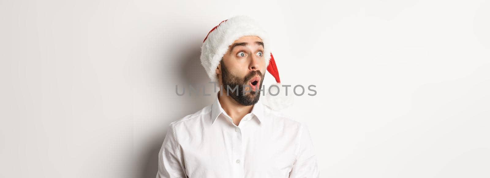 Close-up of amazed bearded man in santa hat, looking left with surprised face, christmas promo offer, standing over white background by Benzoix