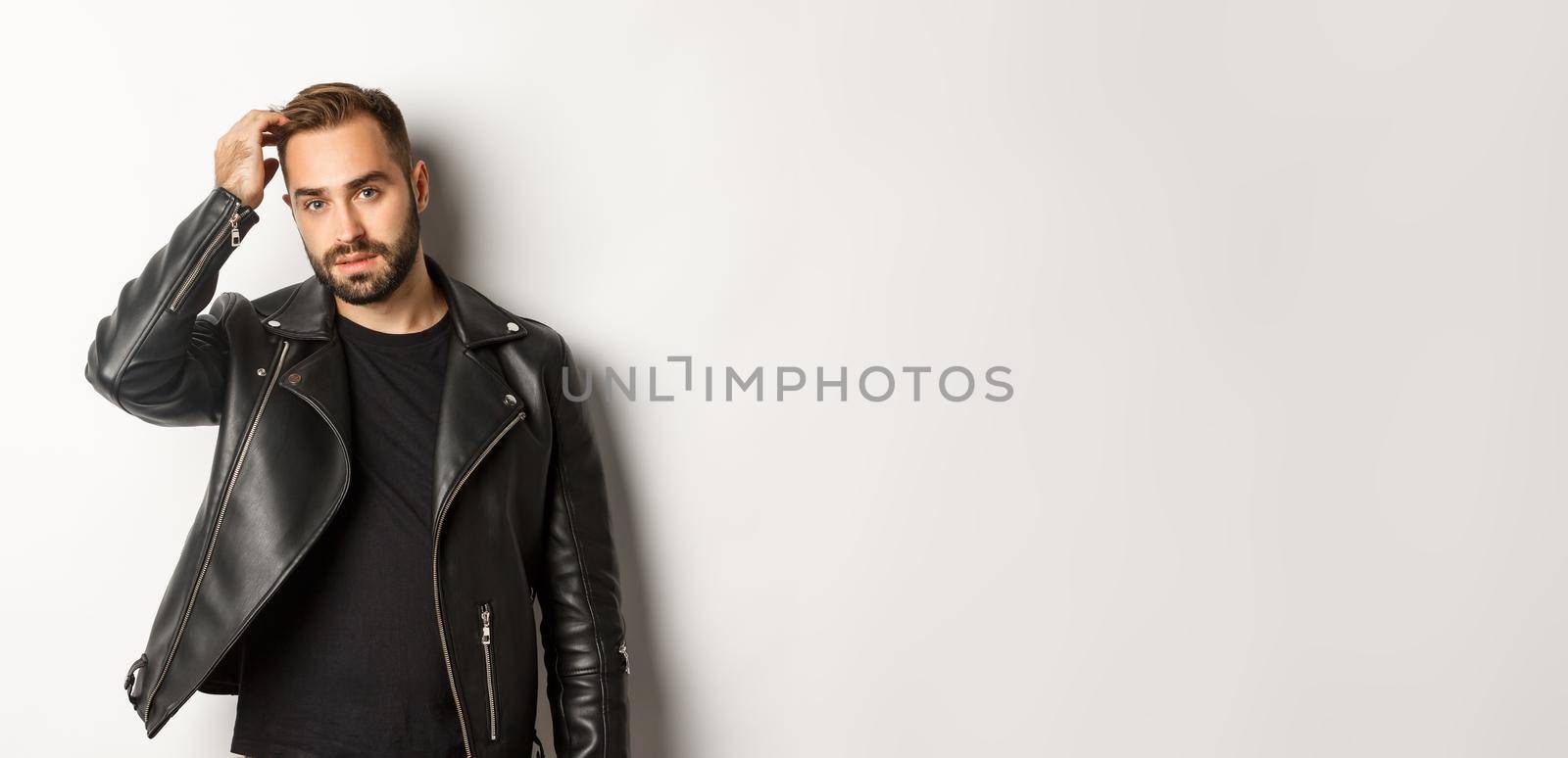 Handsome macho man in black biker jacket, touching his haircut and looking cool, white background.