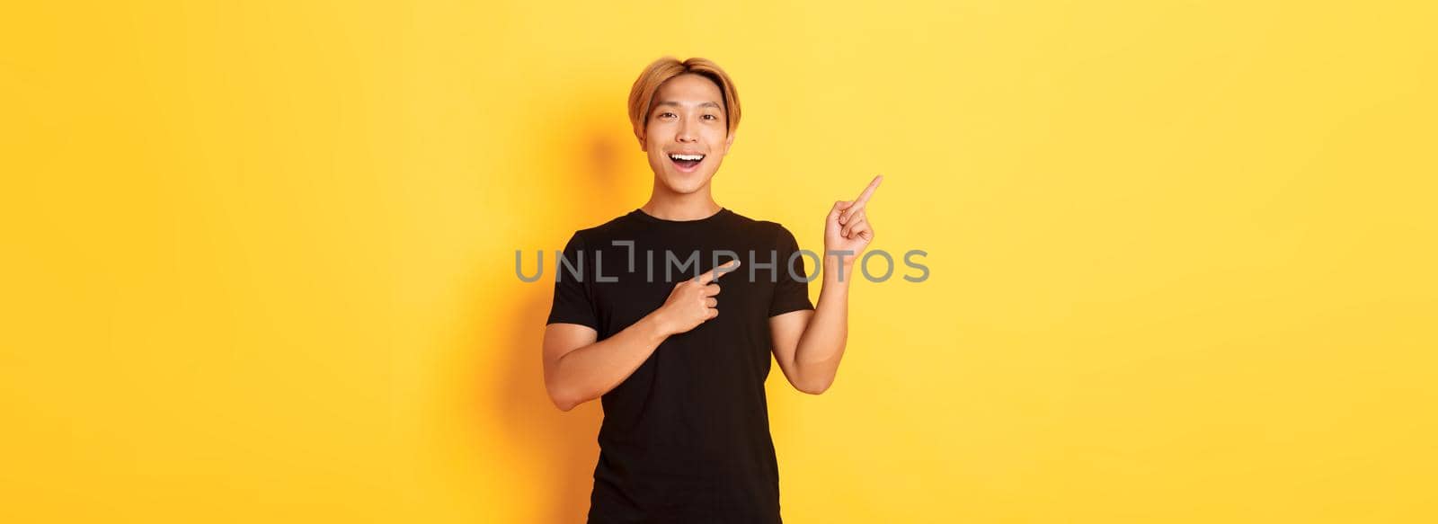 Portrait of cheerful asian man pointing fingers upper left corner, smiling and showing banner satisfied, yellow background by Benzoix