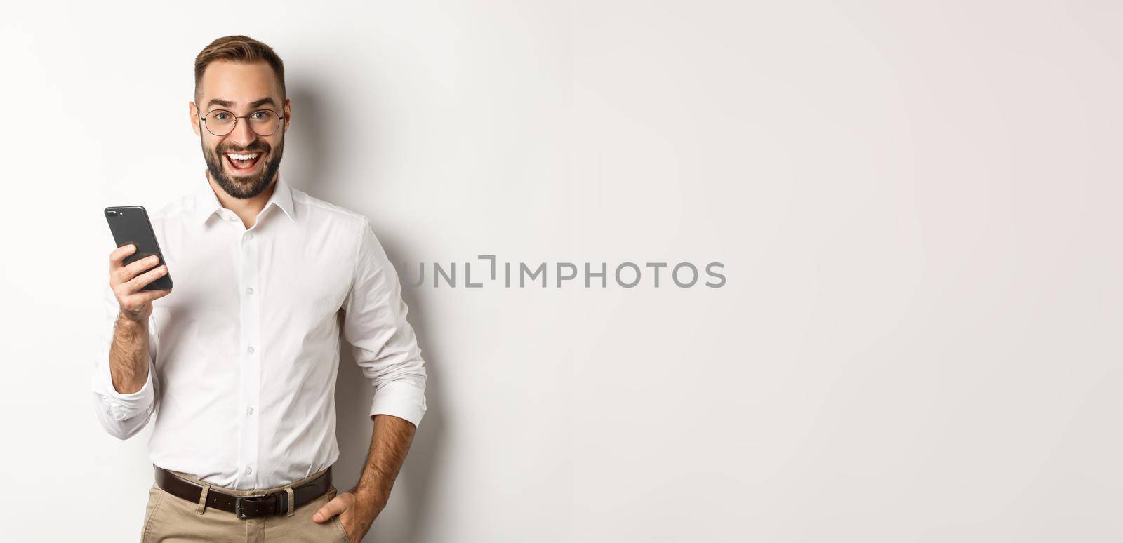 Excited business man using mobile phone, looking amazed, standing over white background.