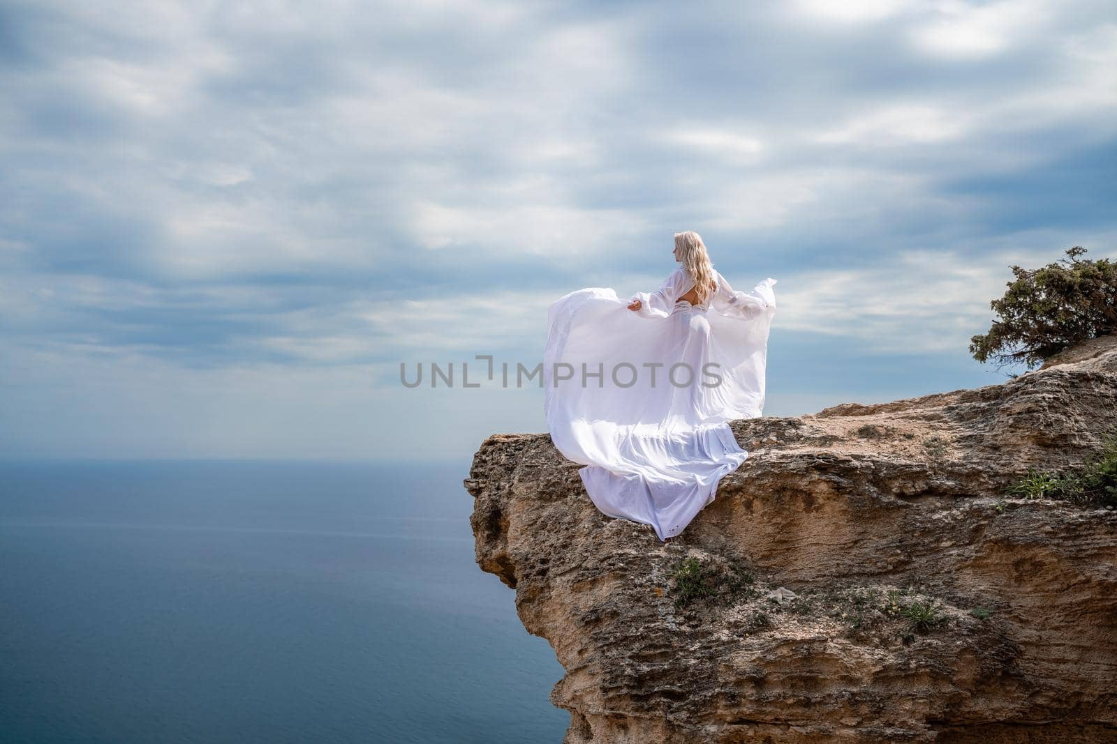 Blonde with long hair on a sunny seashore in a white flowing dress, rear view, silk fabric waving in the wind. Against the backdrop of the blue sky and mountains on the seashore. by Matiunina