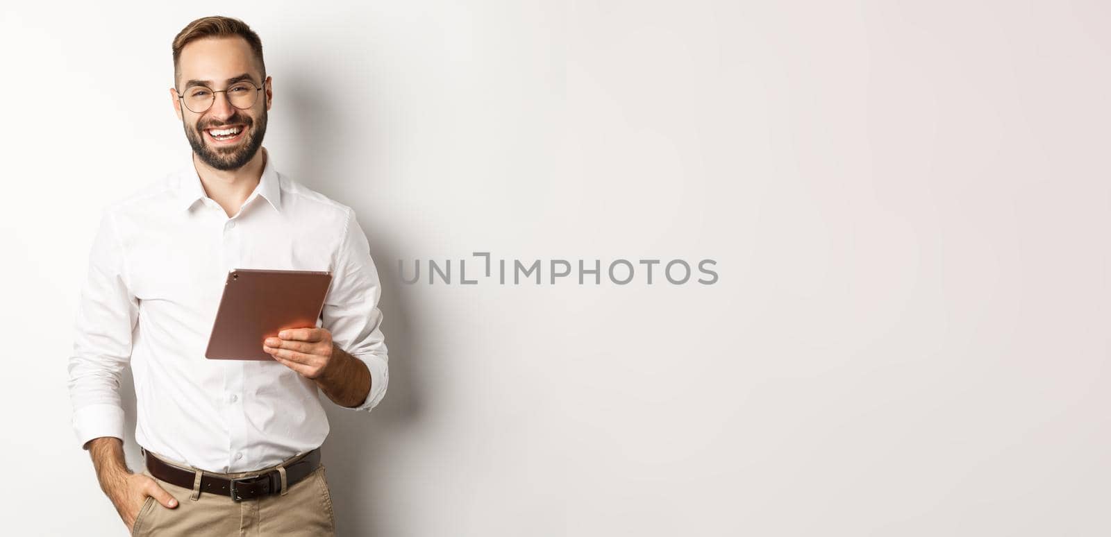 Confident business man holding digital tablet and smiling, standing against white background.