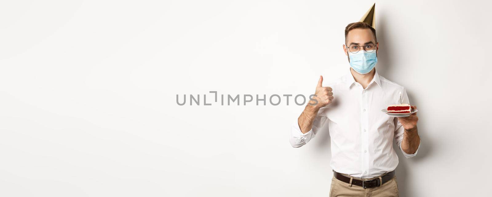 Covid-19, social distancing and celebration. Happy young man in face mask, enjoying birthday party, holding bday cake and make thumb up gesture, white background.