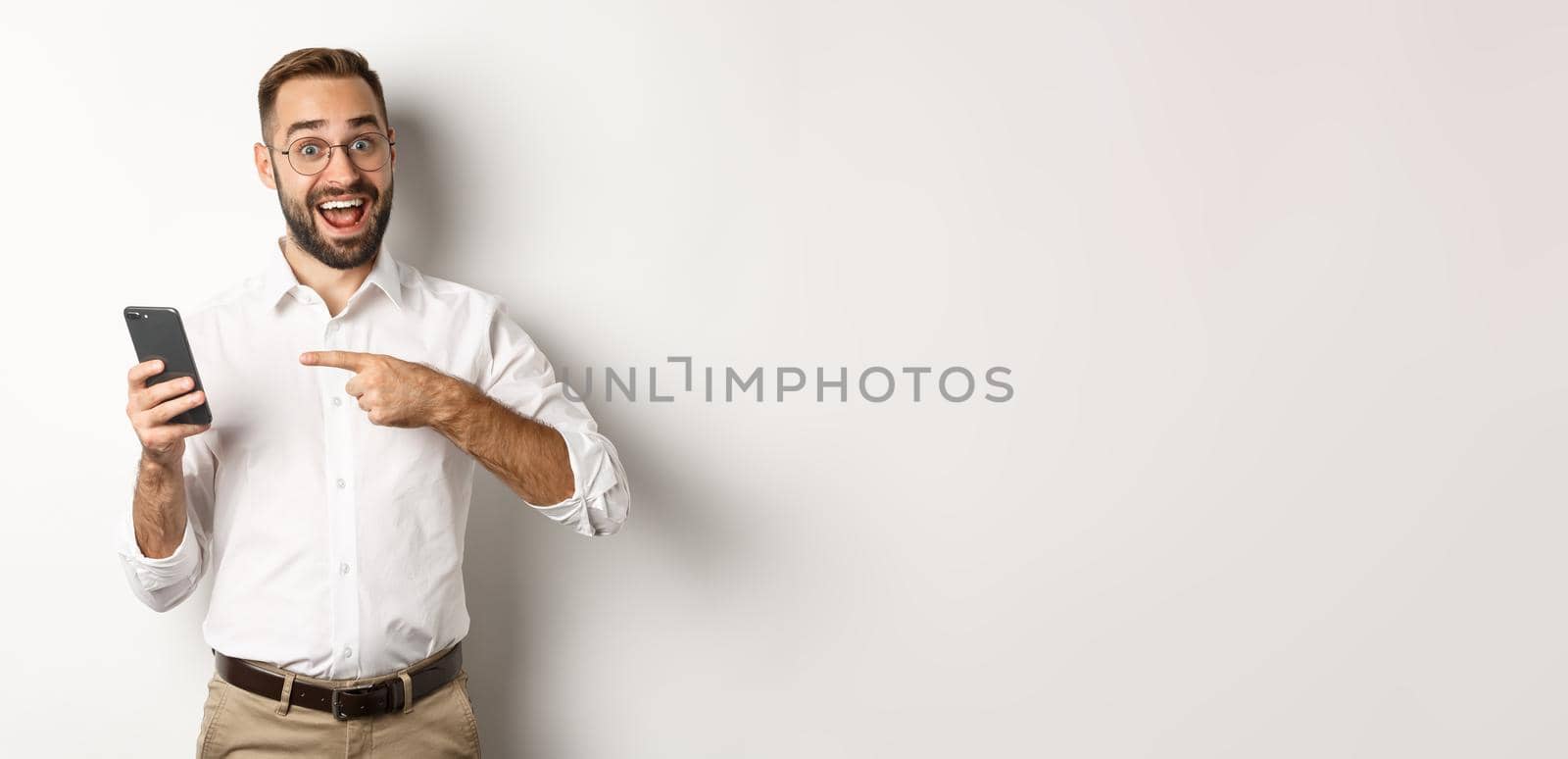 Man looking excited and pointing finger at mobile phone, showing good online offer, standing over white background.